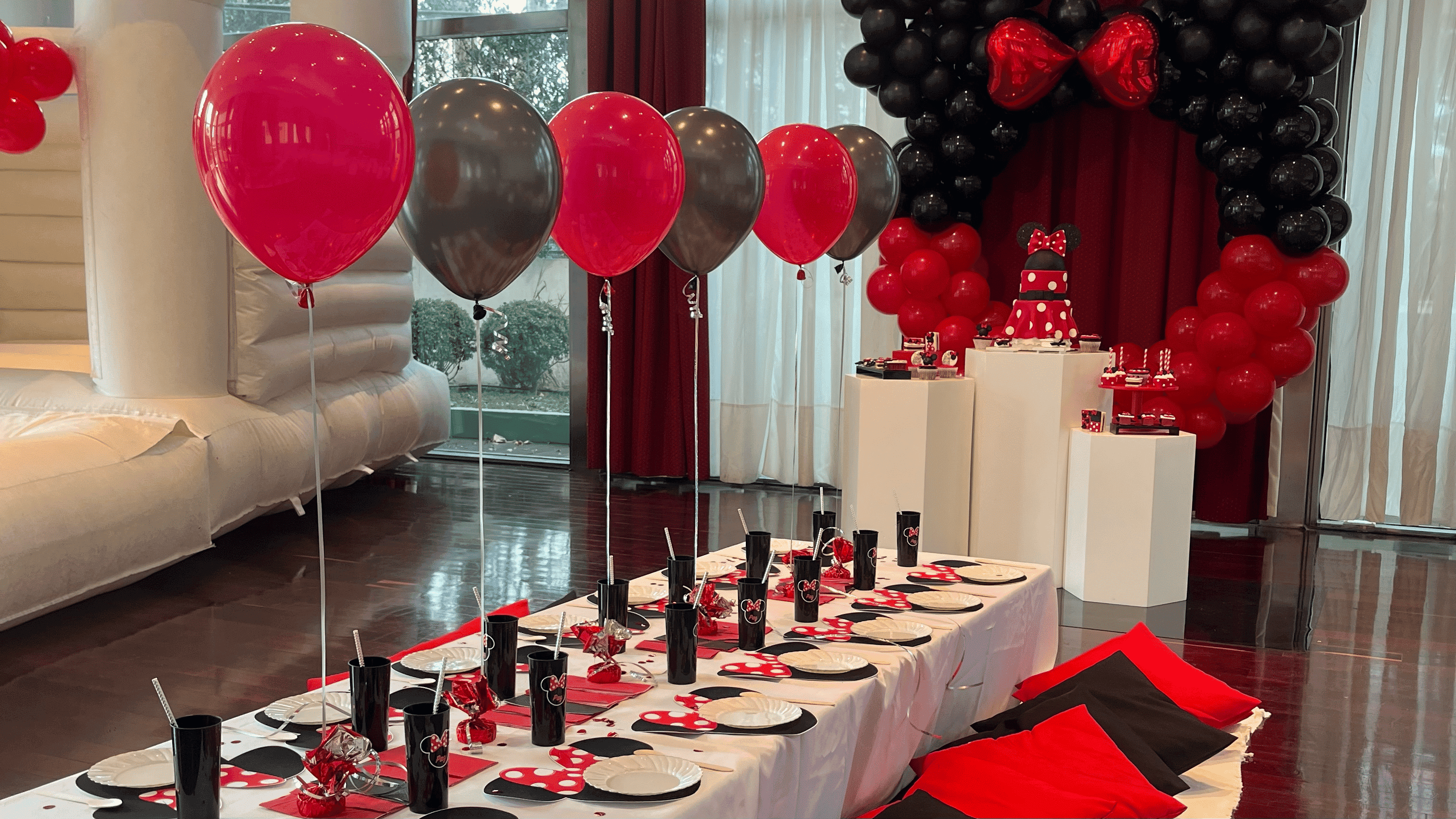 Decoración de cumpleaños infantil. Incluye globos de colores negro, blanco y rojo. Debajo se encuentra una mesa para niños con globos y vajilla personalizada, y al fondo de la imagen se ve una torta y mesa dulce.