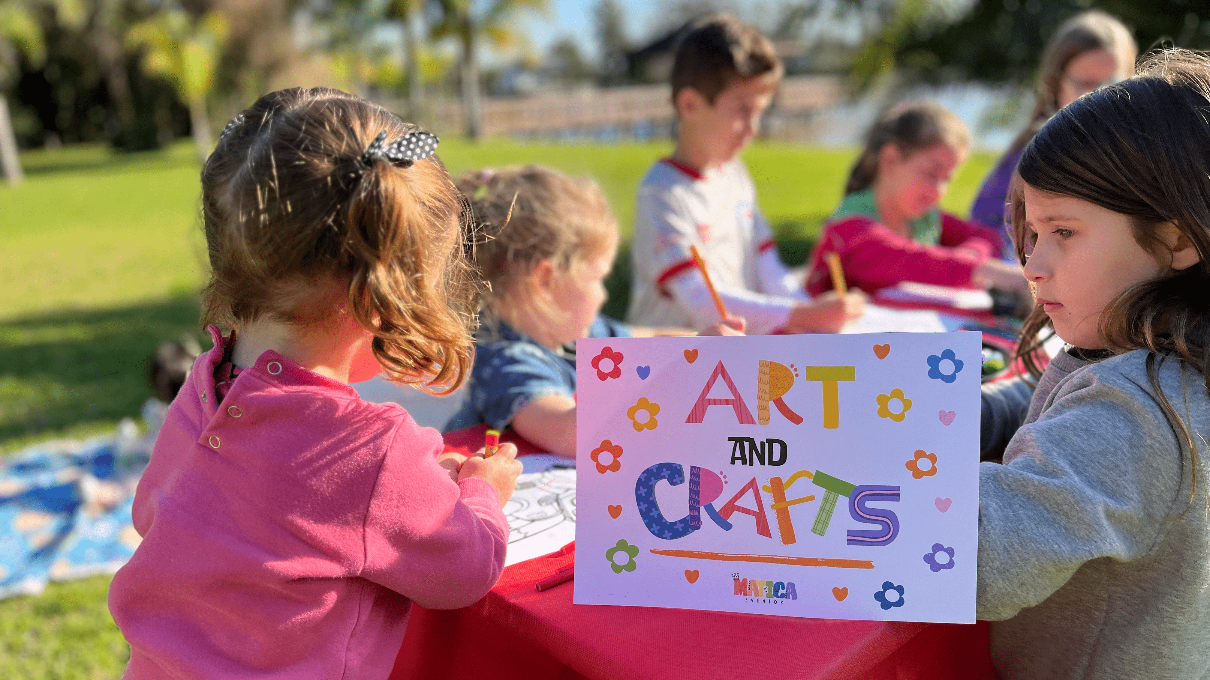 Grupo de niños realizando un taller de pintura en una mesa. En la foto aparece un cartel con el texto 'Arts & Crafts'.