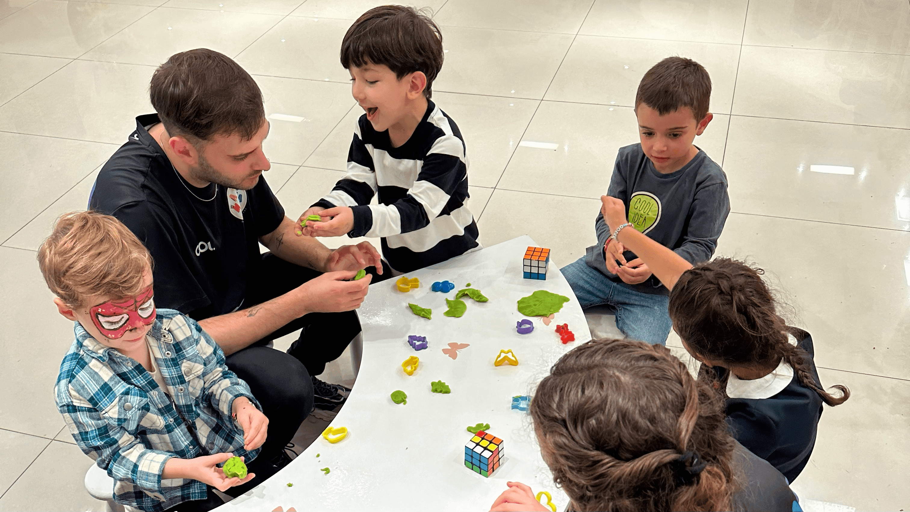 Grupo de niños sentados en una mesa con juguetes y plastilina. El animador de Matica Eventos está a la izquierda de la foto, jugadando y cuidando a los niños.