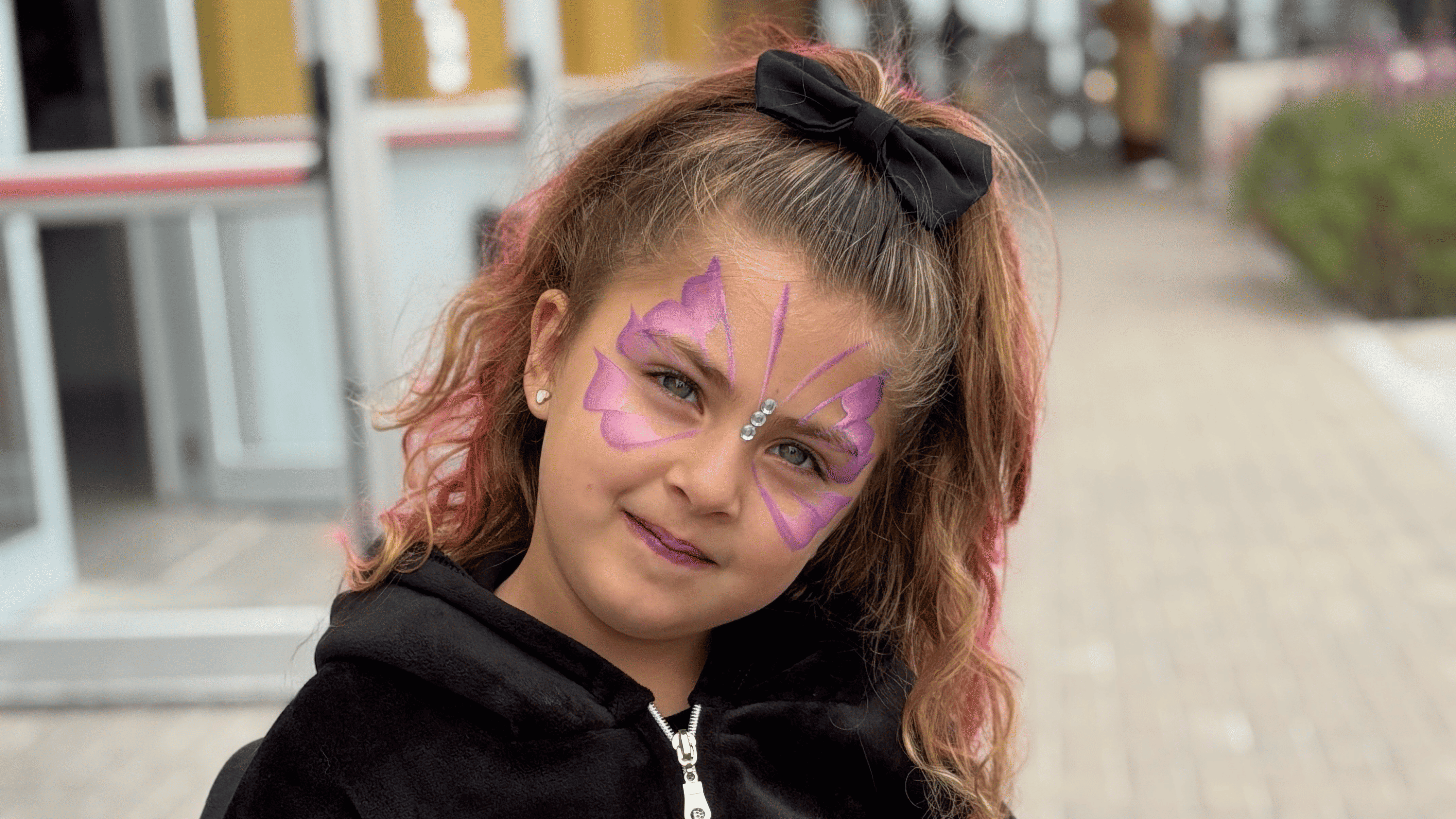 Niña con maquillaje artístico en la cara. El dibujo es una mariposa rosa con brillos.